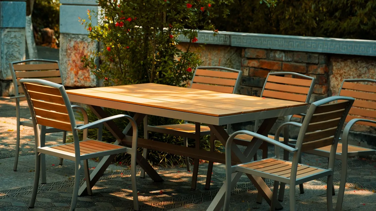 A rectangular wooden-topped patio table with metal-framed slatted chairs beside a garden wall with red flowers in dappled sun