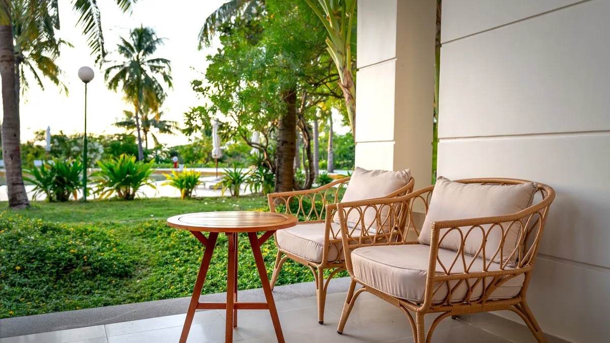 Two cushioned woven armchairs and a small round wooden table on a covered patio looking out over a garden with palm trees