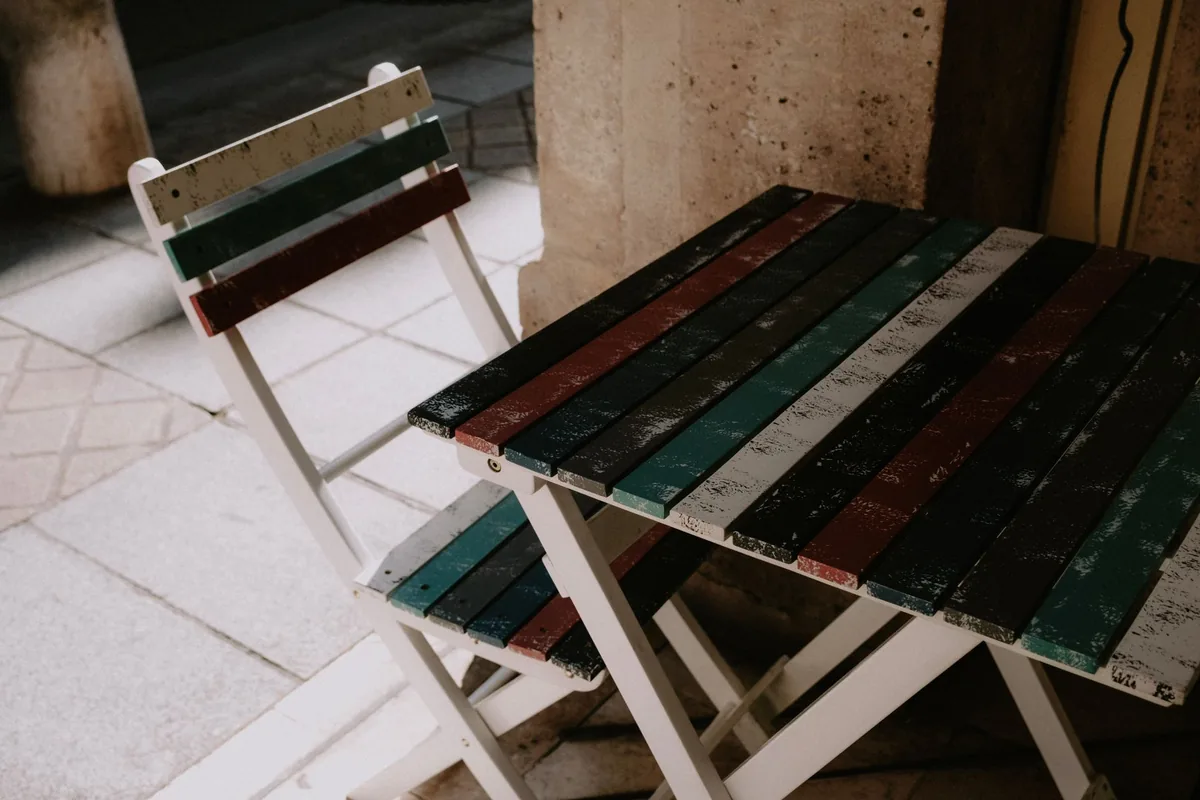 A folding wooden table and chair with painted slats standing on pale paving slabs beside a stone wall