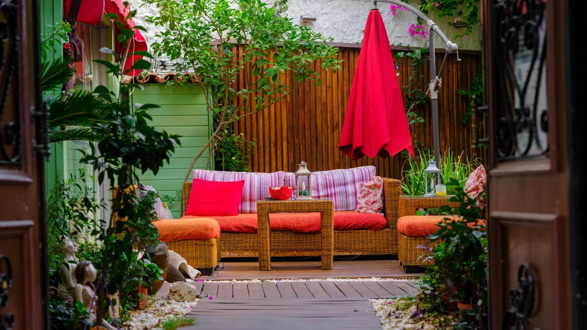 A garden patio with a woven sofa and armchairs with orange and pink cushions, a closed red umbrella, potted plants and a wooden fence behind