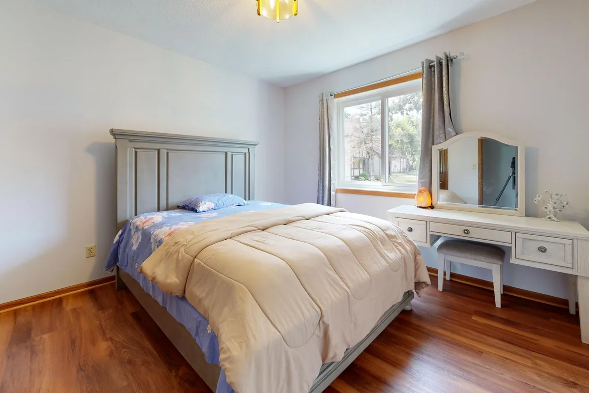 A bedroom with a made bed, a grey headboard, a window with curtains and a white dressing table with a mirror and stool