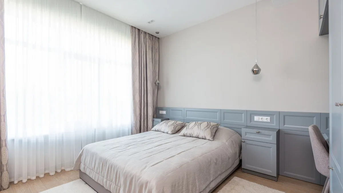 A small bedroom with a made bed, grey built-in cabinets along the wall, sheer curtains at a tall window and a desk chair at the right edge