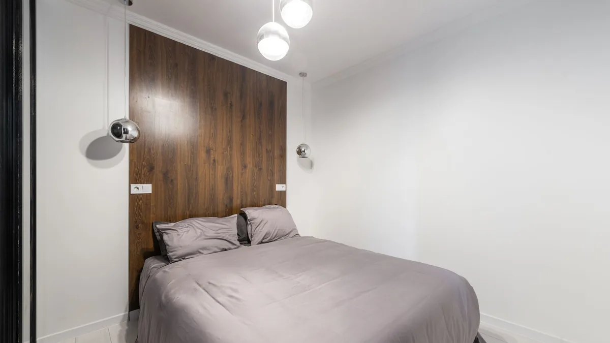 A small bedroom with a bed fitted between two walls, a wood-panel headboard wall and pendant lights