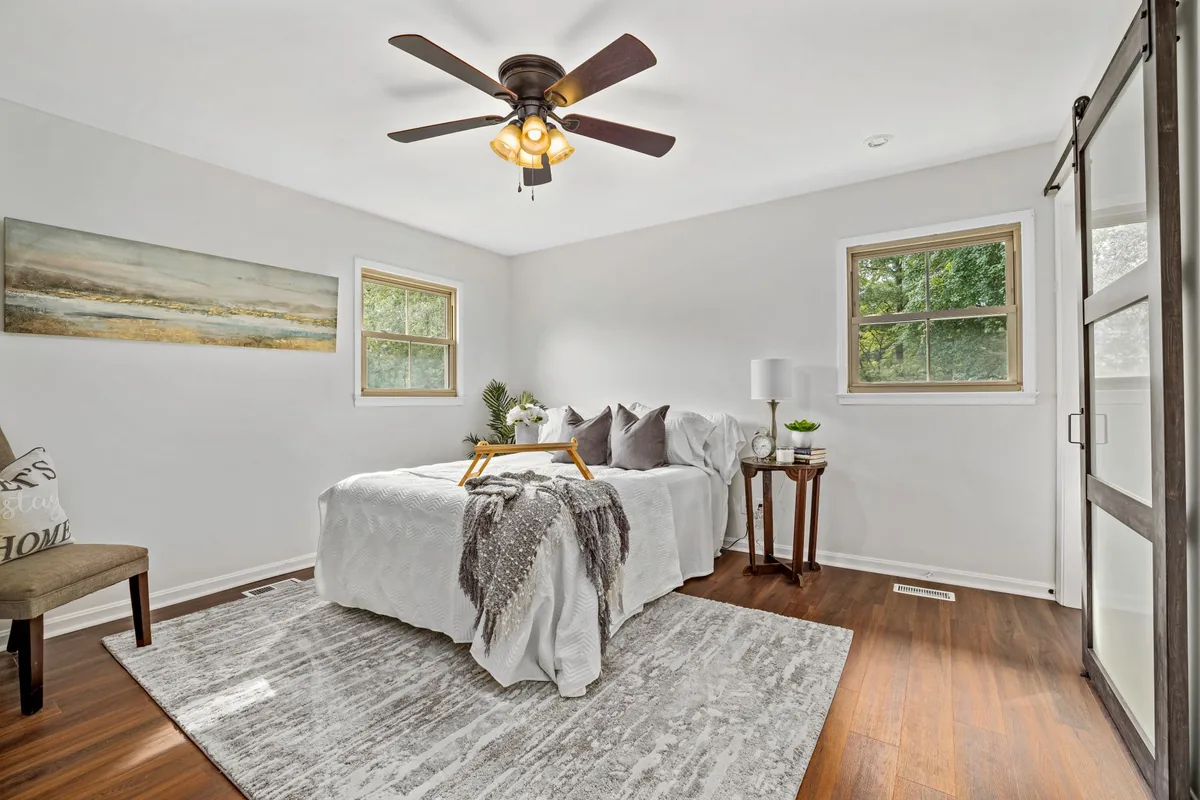 A bed with white bedding and a grey throw on a wood floor, a large grey patterned rug under its lower half and a round wooden side table on the bare floor beside the headboard