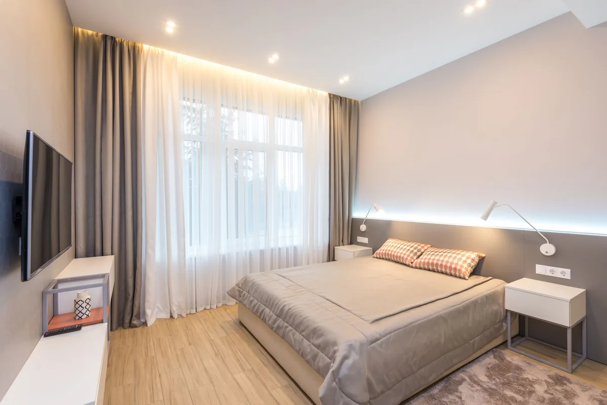 A bedroom with a large bed between two white nightstands, a tall window with curtains and a grey rug showing at the foot of the bed on a light wood floor
