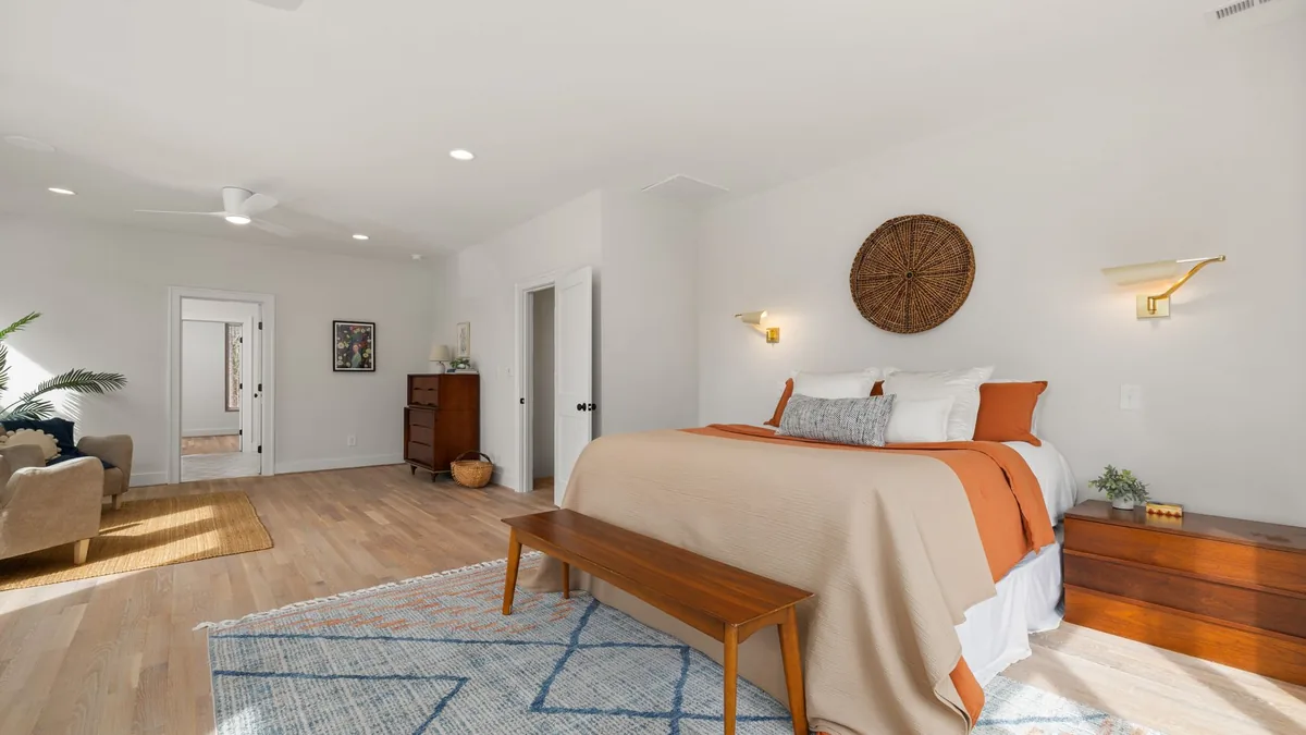 A large bedroom with a king-size bed in neutral bedding, a wooden bench at its foot and a blue and cream patterned rug under the lower part of the bed, with a wooden nightstand on the bare floor