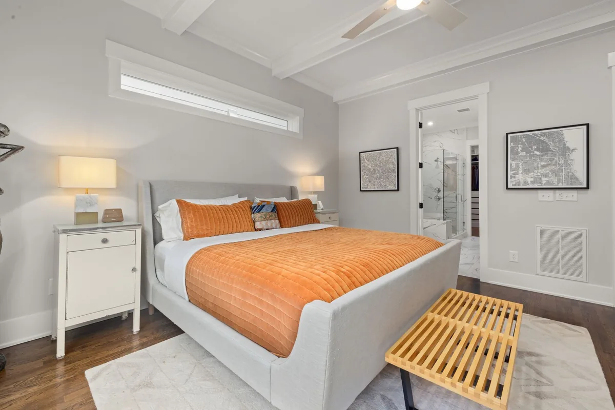 A bed with an orange quilt and a slatted wooden bench at its foot, both on a pale patterned rug, with a white nightstand on the wood floor beside the headboard