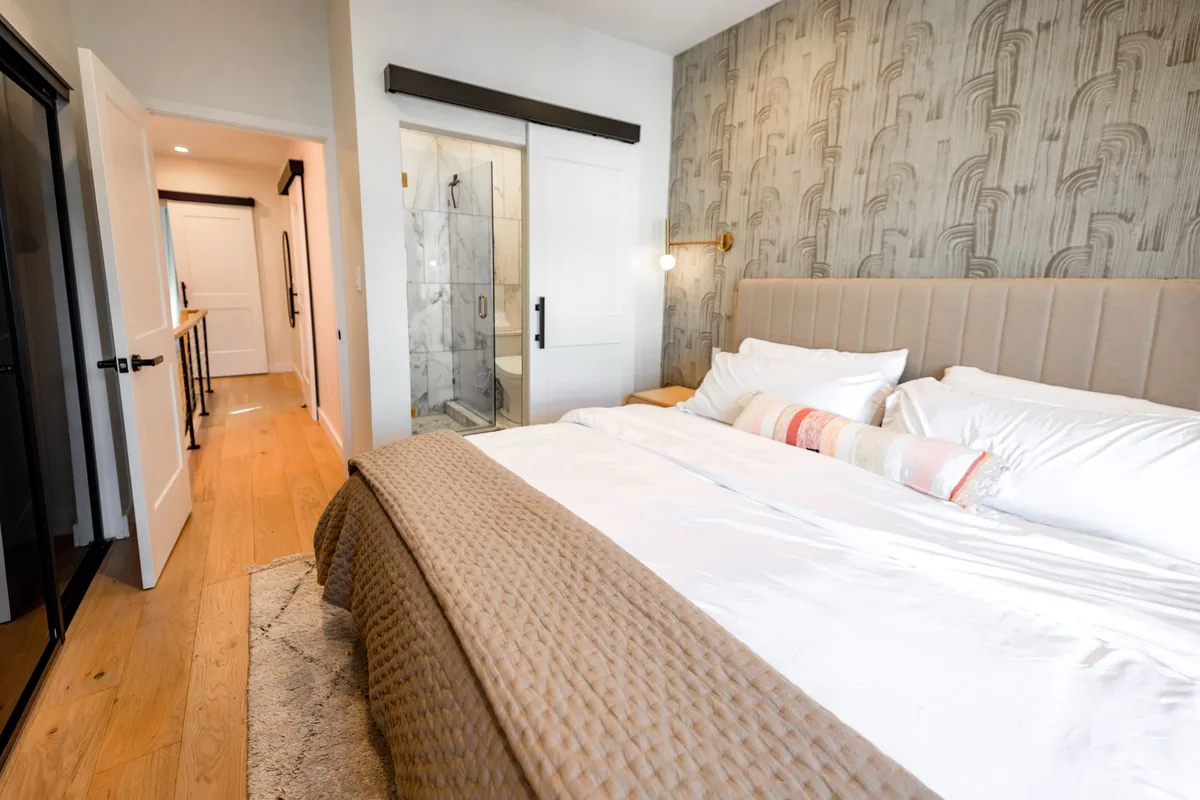 A narrow bedroom with a large bed against a patterned wall, a small shaggy rug on the wood floor beside it and an open door to the hallway
