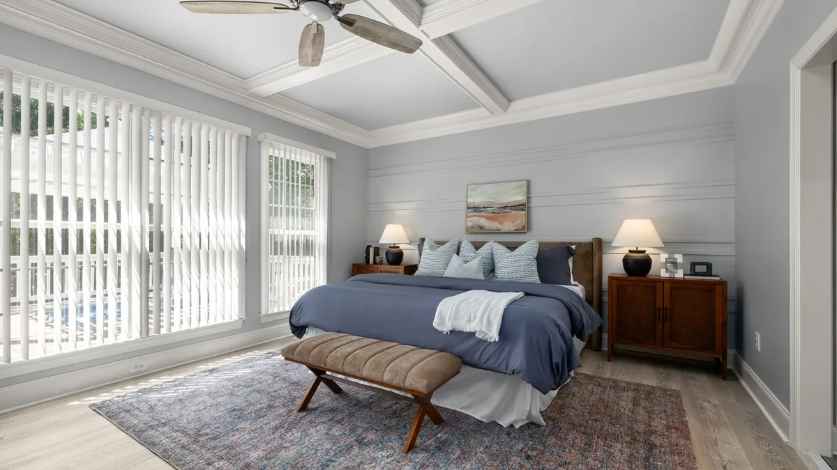 A bedroom with a bed in navy bedding, an upholstered bench at its foot and a faded red and blue rug under the lower part of the bed, with wooden nightstands on the bare floor