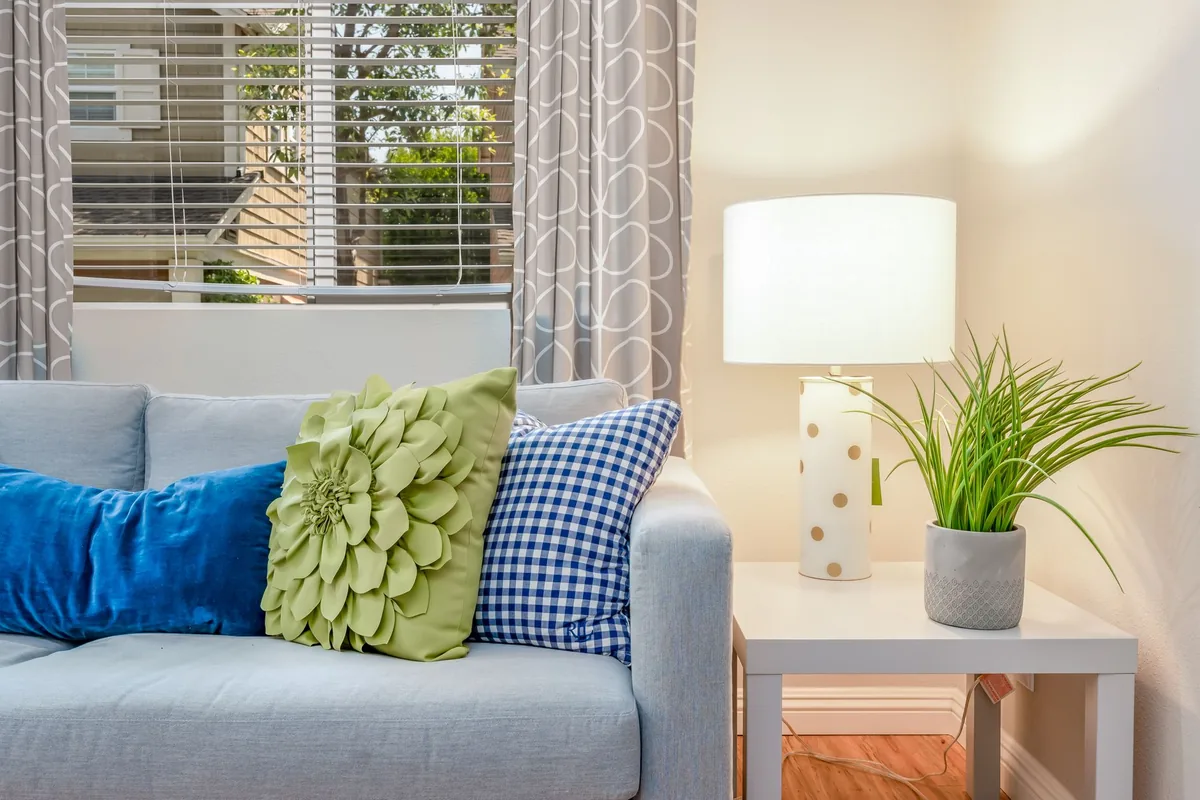 A light grey sofa with colourful cushions beside a white square side table holding a table lamp and a potted grass plant