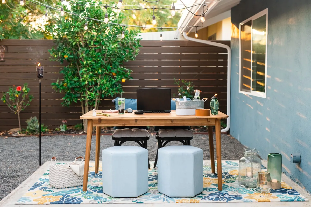 A small backyard patio with a wooden table, two pale upholstered stools on a patterned outdoor rug and string lights overhead