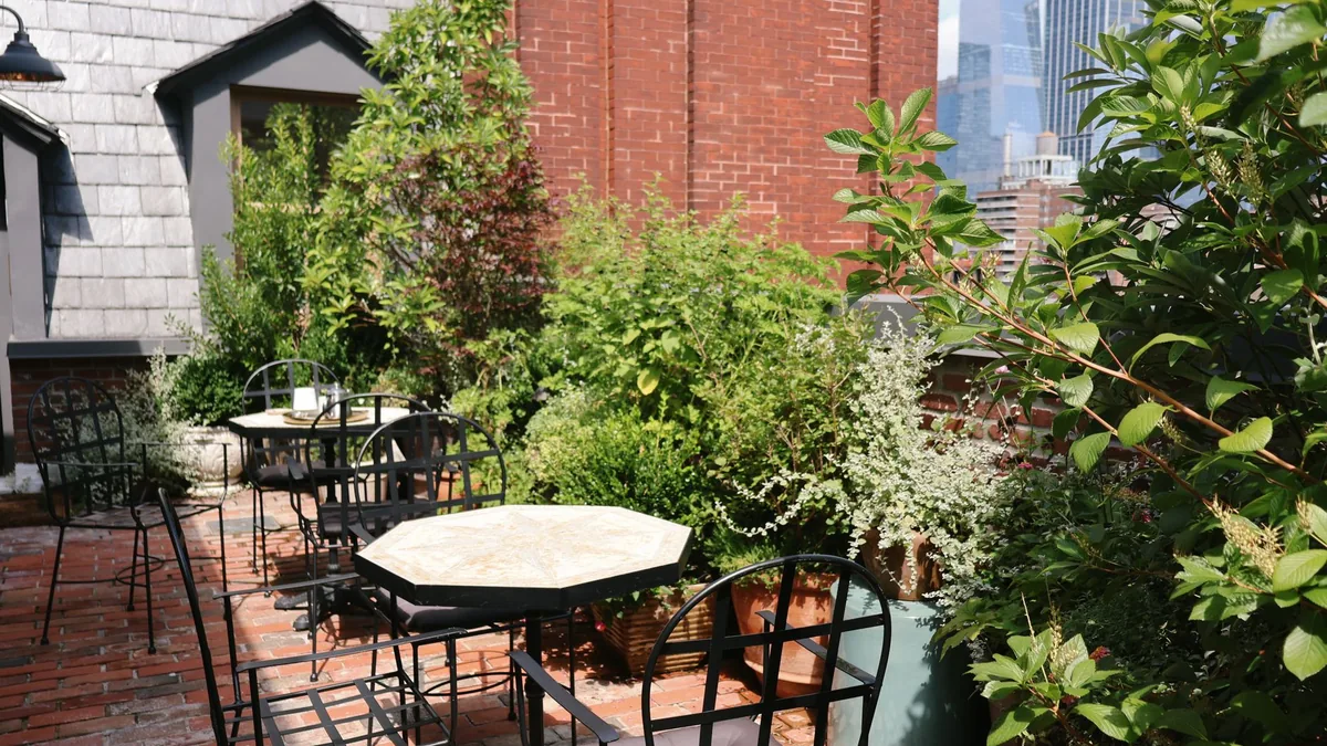 A small brick-paved roof terrace with black metal bistro tables and chairs surrounded by potted shrubs and climbing plants