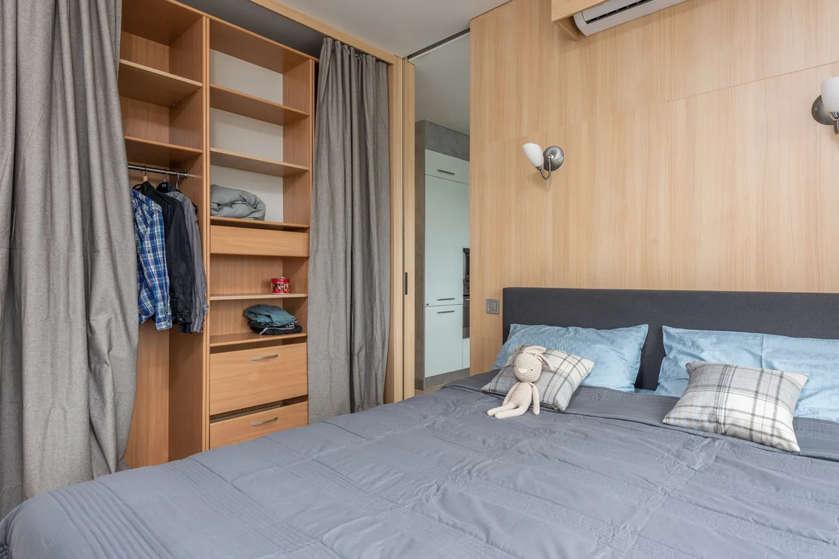 A small bedroom with a double bed beside an open cupboard of shelves, drawers and a short hanging rail behind a gray curtain