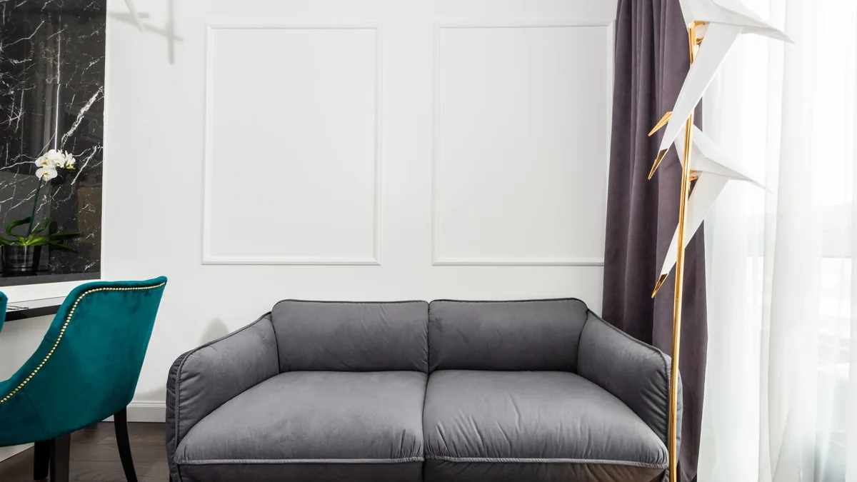 A grey two-seat sofa with plump cushions against a white panelled wall, between a teal velvet chair and a long curtain