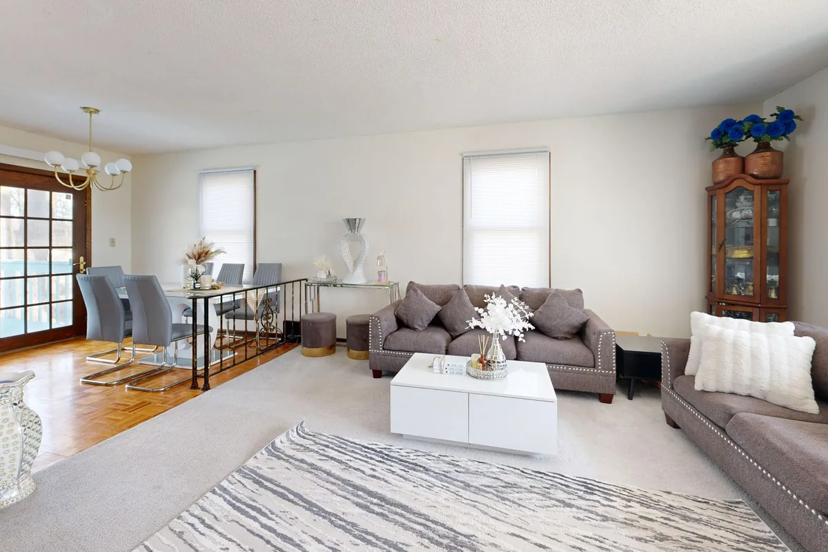 A bright living room with two grey sofas around a white coffee table on a patterned rug, with a dining table beyond