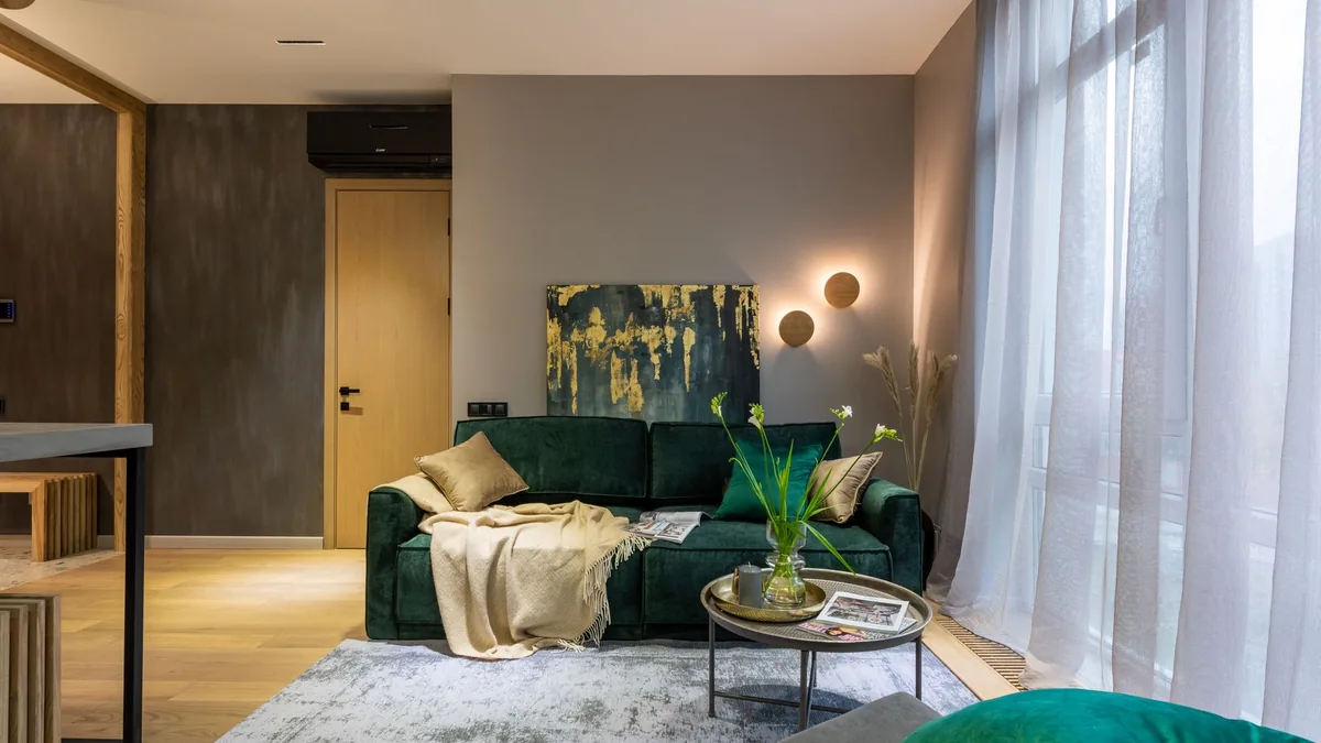 A small living room with a dark green two-seat sofa, a round tray table on a grey rug, a wooden door and sheer curtains beside a tall window