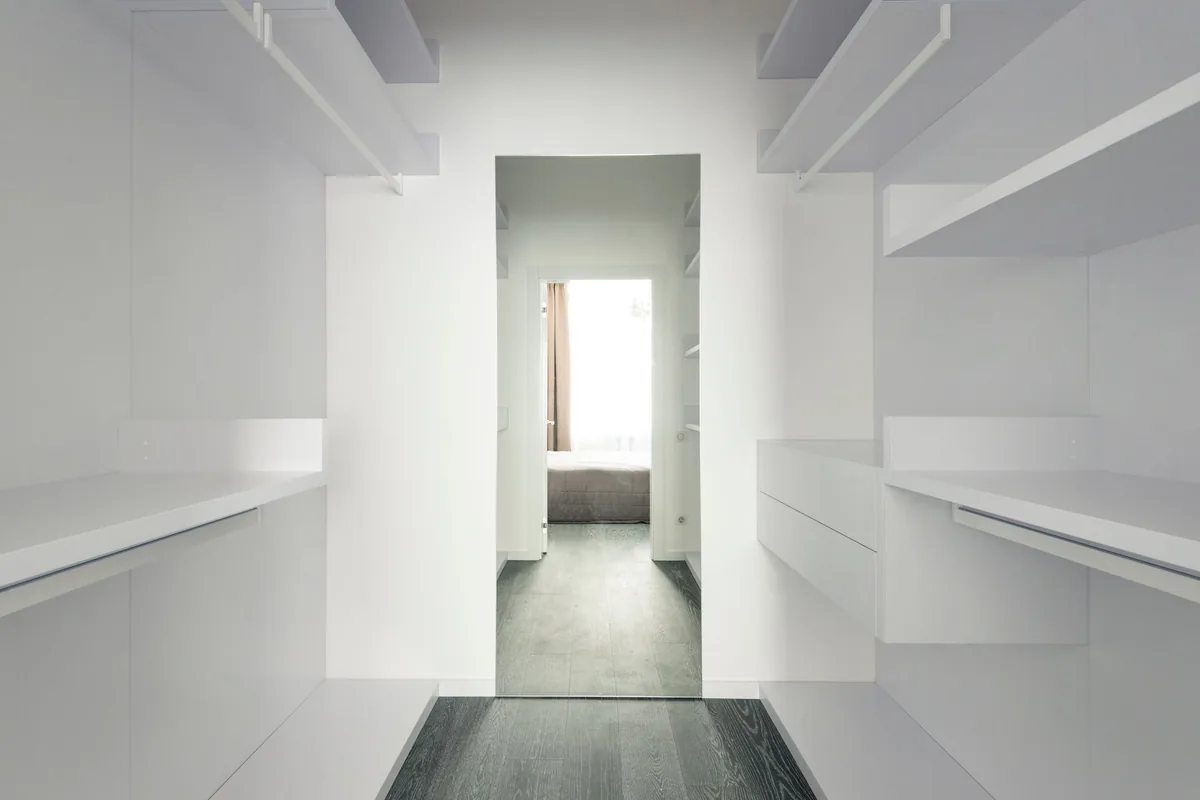 An empty white walk-in closet with shelves and rails on both long walls and a door at the far end