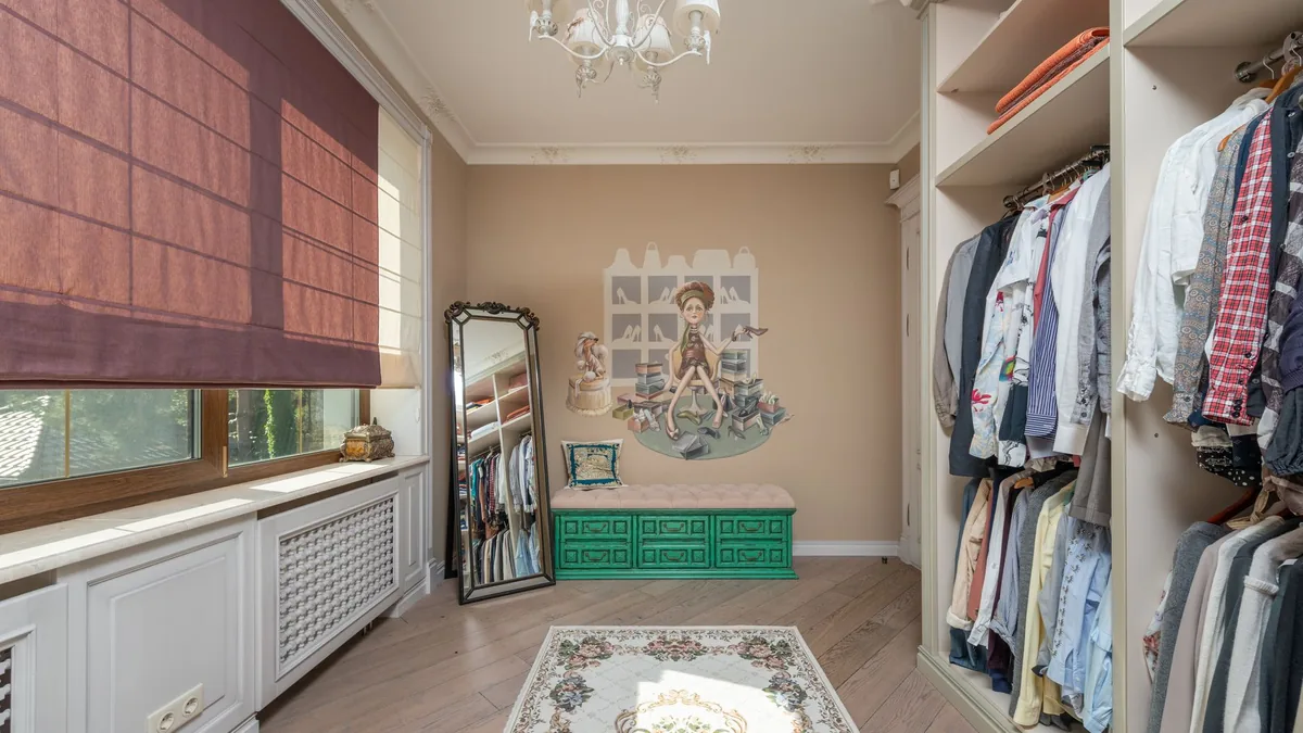 A walk-in dressing room with clothes on open rails, a tall mirror, a green bench and a window with a red blind